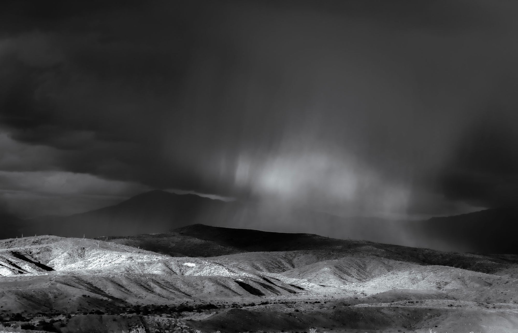 Black and white photograph of dramatic rain shafts descending over rolling hills in Tonto National Forest, Arizona